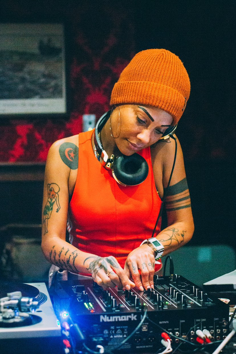 Woman DJ wearing orange beanie and red top, adjusting equipment in a dimly lit studio.