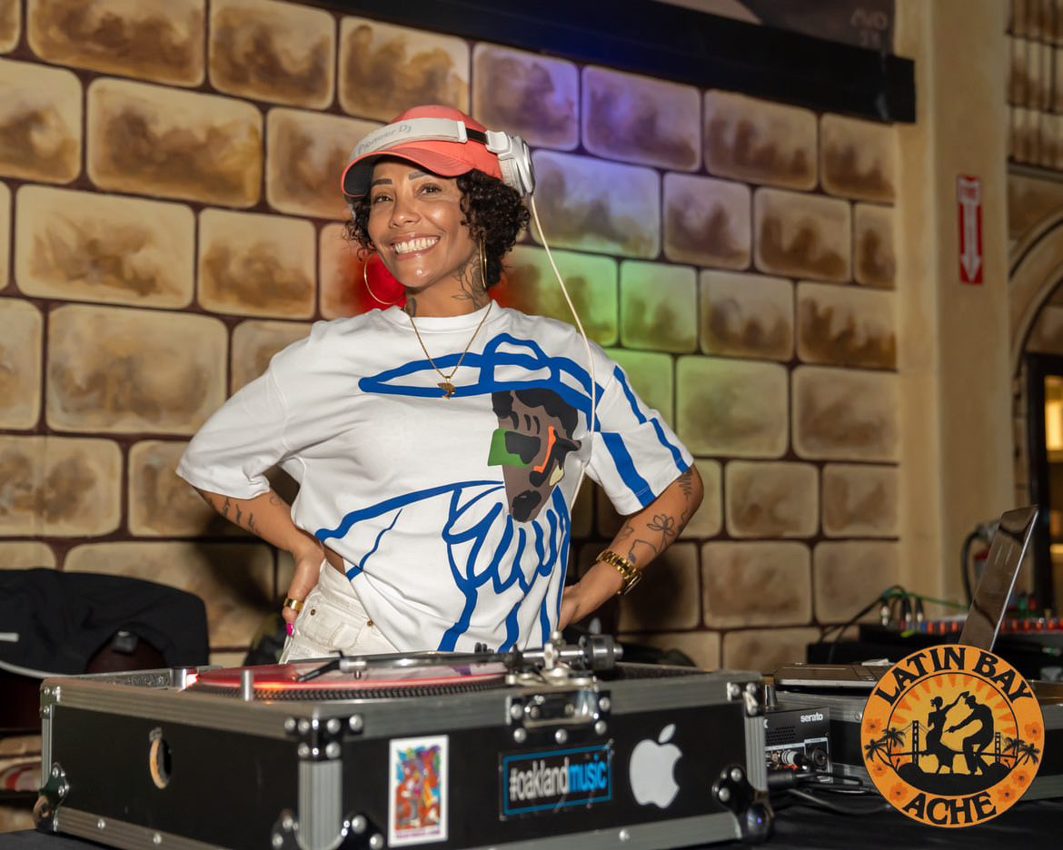 Woman DJ wearing a white shirt, red hat, and headphones, smiling behind DJ equipment with a brick wall background.
