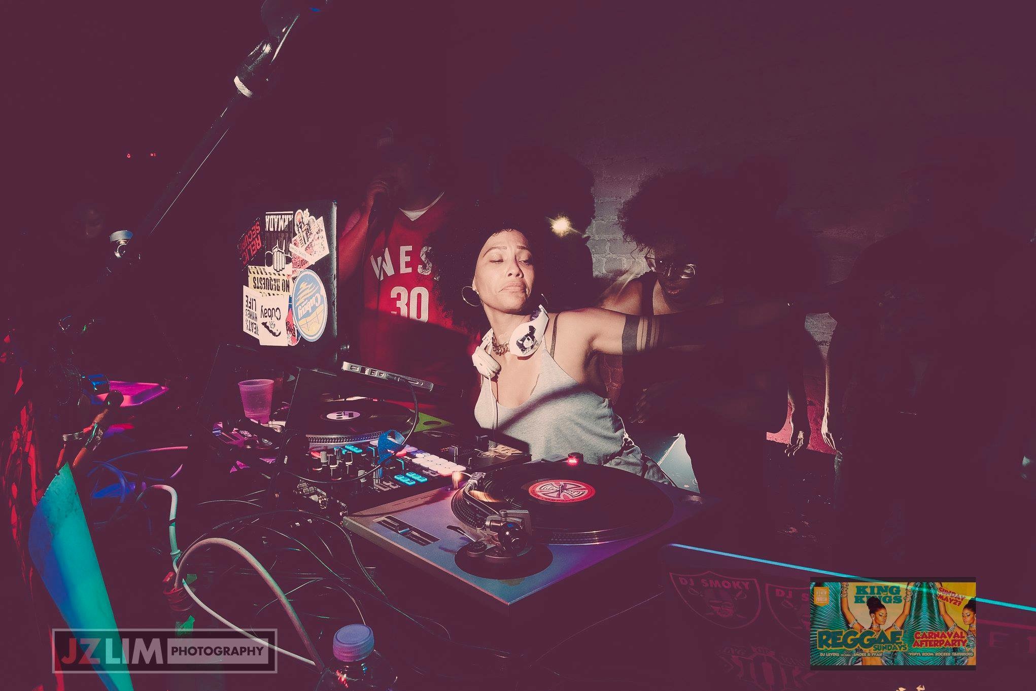 DJ woman with headphones performing at a booth, with equipment and a dark background.
