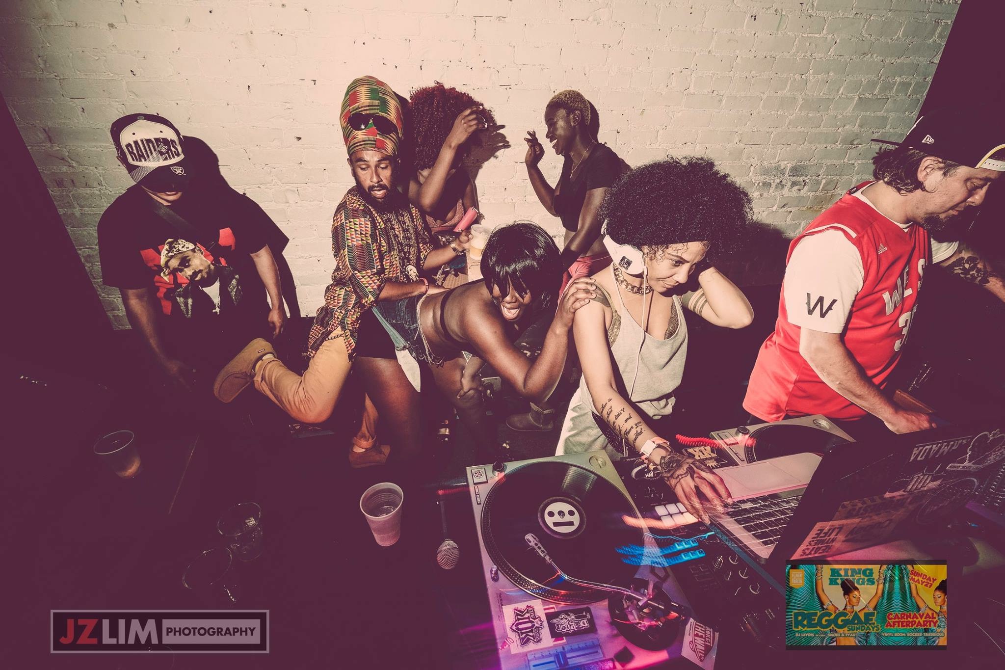 Group of people dancing and DJing in a dimly lit indoor space, some wearing hats and casual clothing.