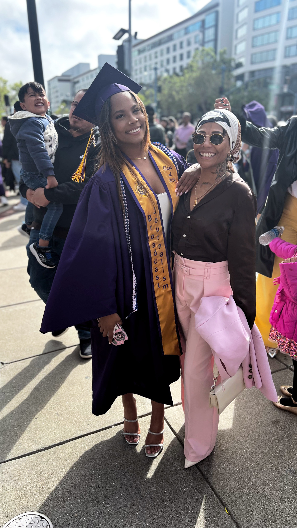 Two women smiling at a graduation event, one in a cap and gown, the other in casual attire, outdoors.