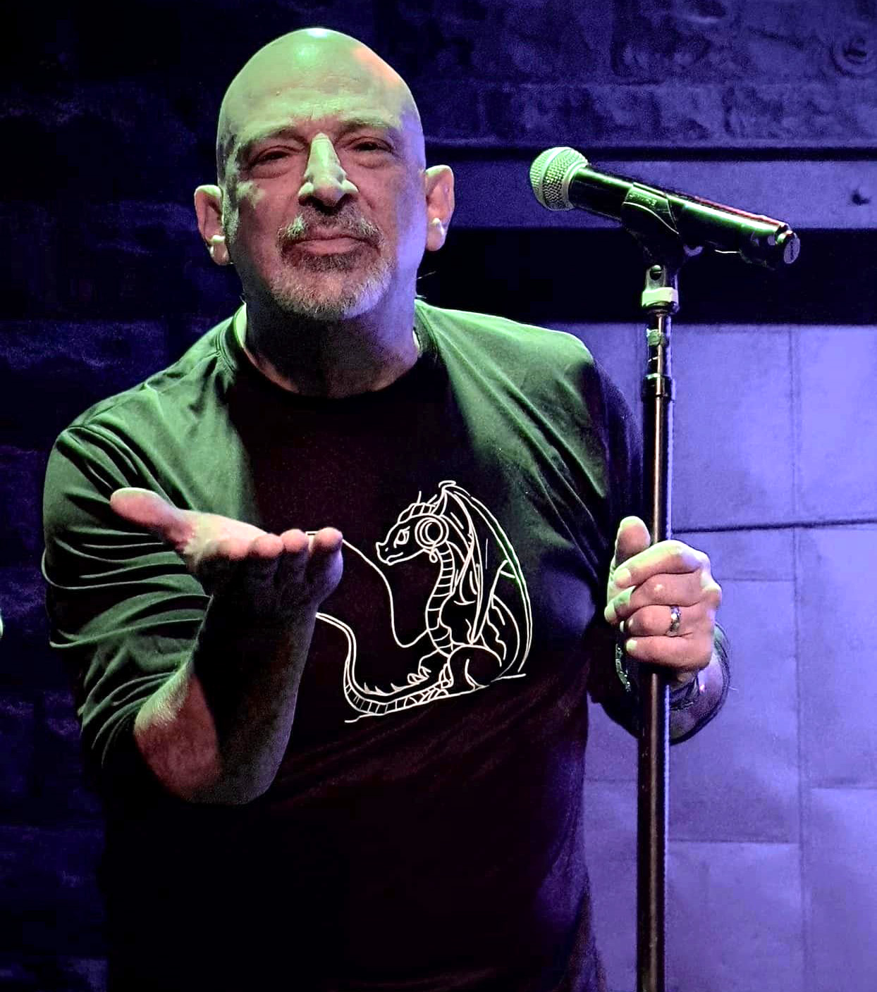 Bald man with beard on stage, holding microphone, wearing a black shirt with a white unicorn design, gesturing with his hand.