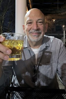 Smiling man holding a glass of beer at an outdoor setting at night.