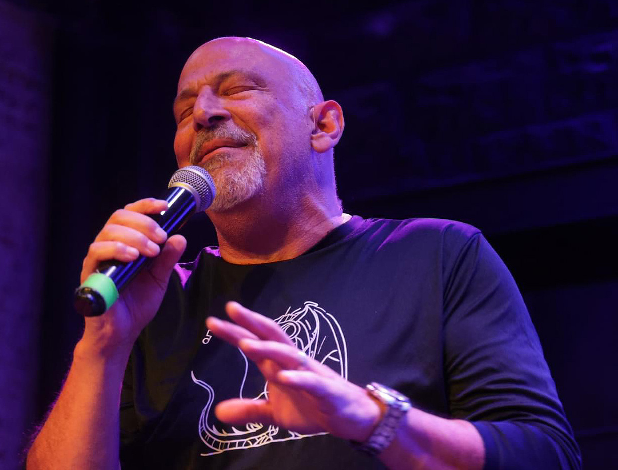 Bald man singing into a microphone with eyes closed, wearing a dark shirt, on stage with dark background.
