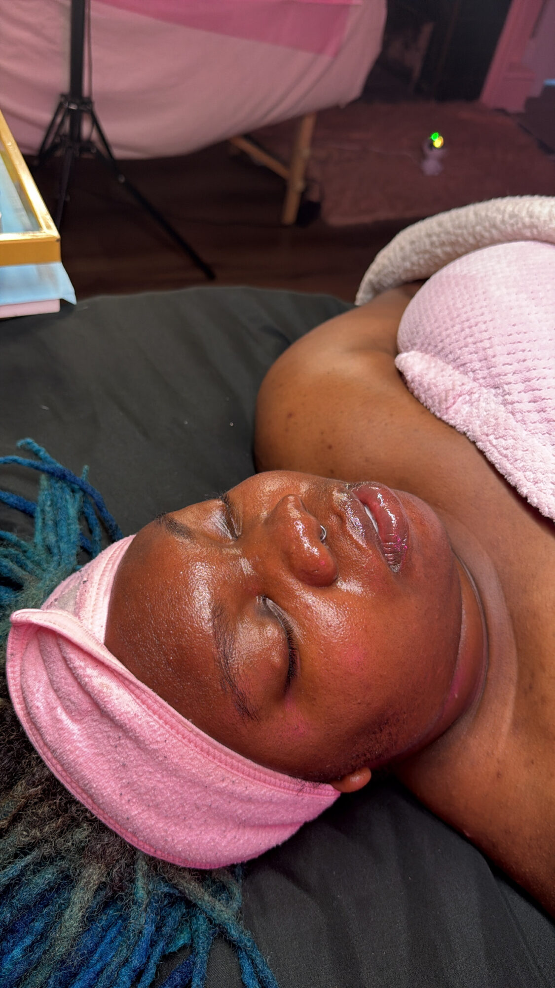 Baby with pink headband and towel resting on a black surface, eyes closed, in a relaxed state.