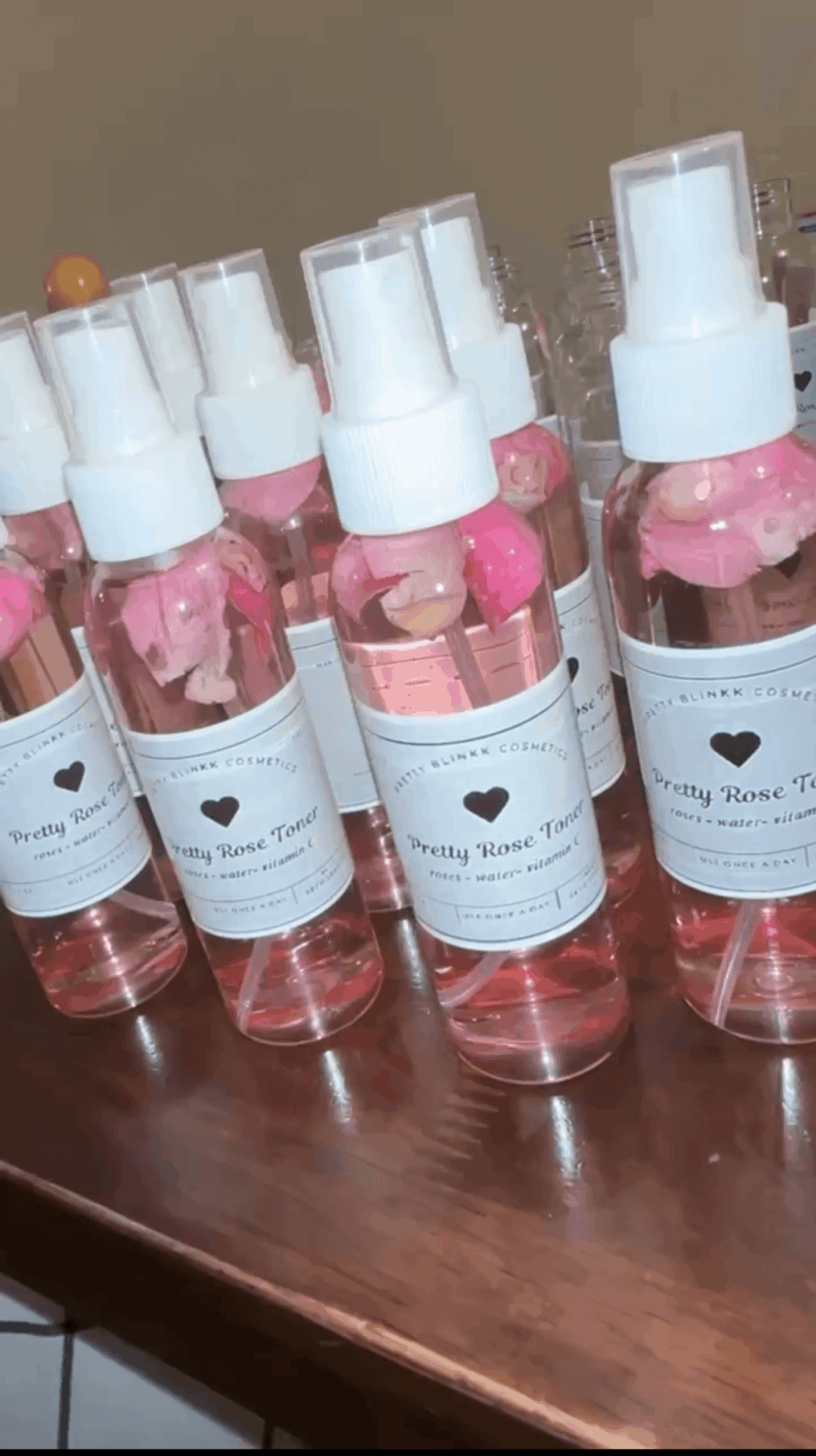 Six small spray bottles with pink liquid and pink flowers inside, placed on a wooden surface.