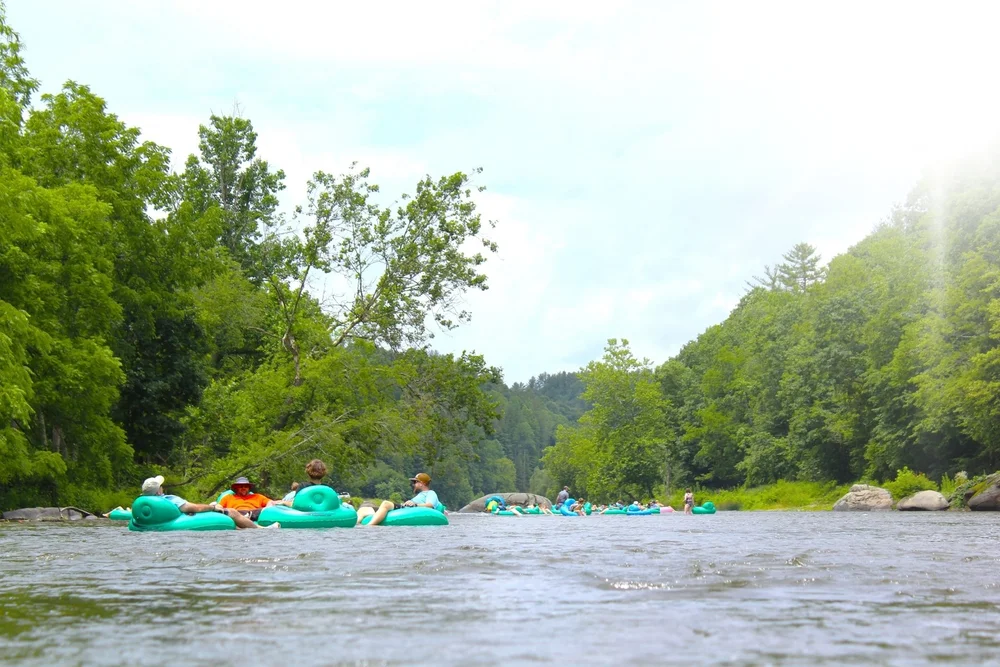 People floating on inflatable tubes in a river surrounded by green trees, with sunlight shining from the top right corner.