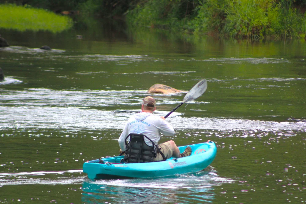 Person kayaking on a river with green trees in the background, wearing a white shirt and beige shorts, paddling with a double-bladed paddle.