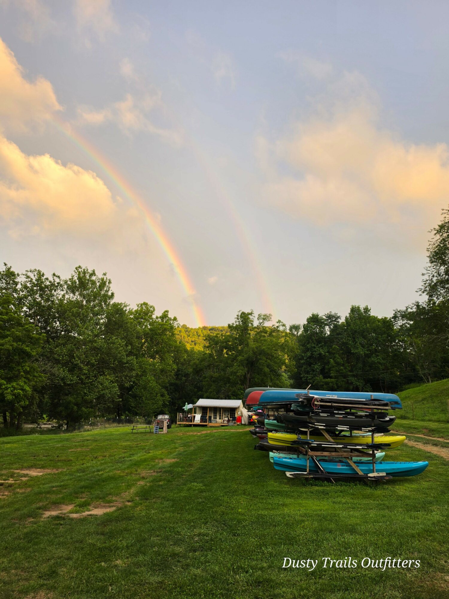 Rainbow over a grassy area with paddleboards and kayaks, trees, and a cloudy sky.
