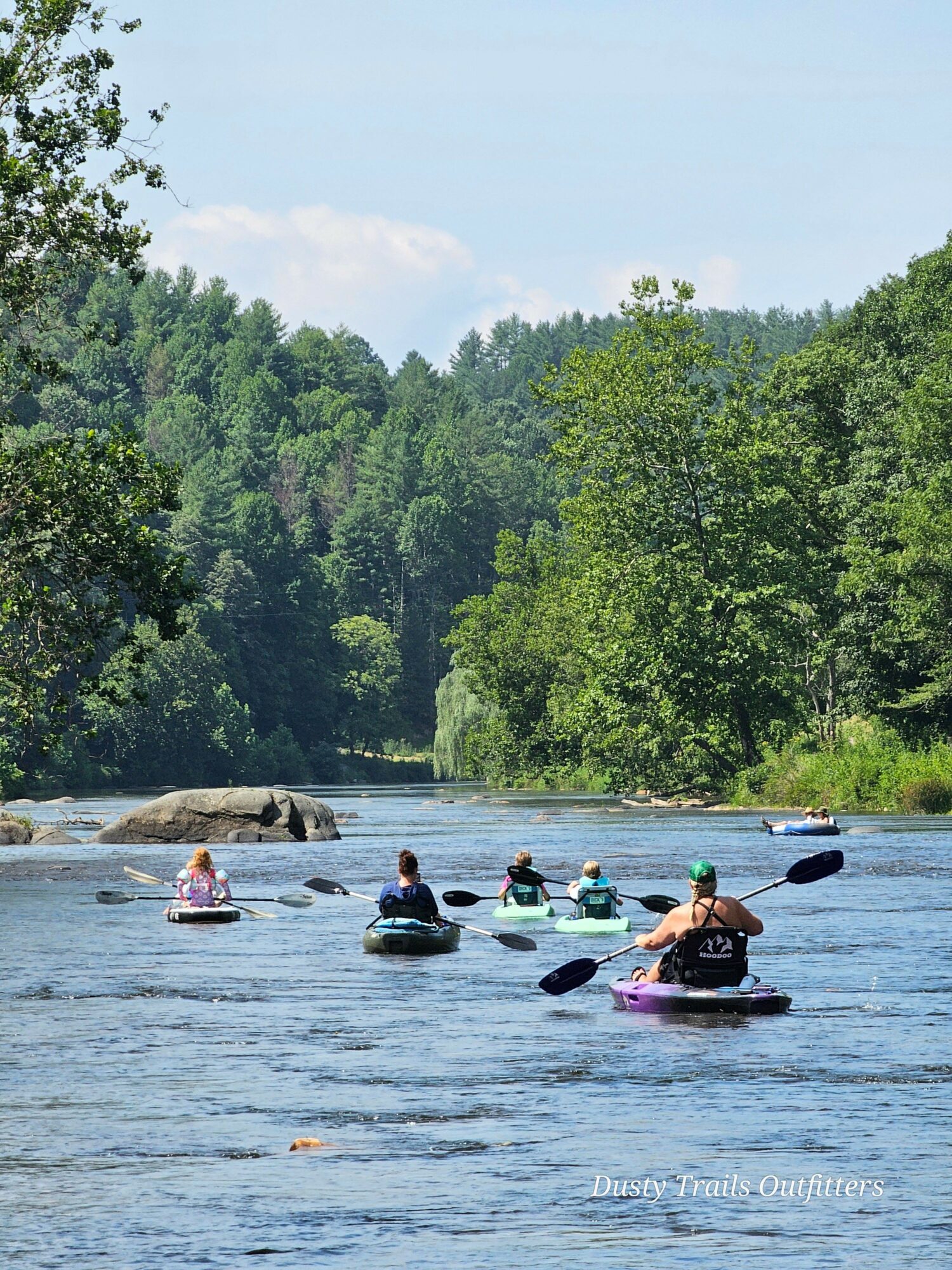 People paddleboarding on a river with lush green trees in the background, under a partly cloudy sky.