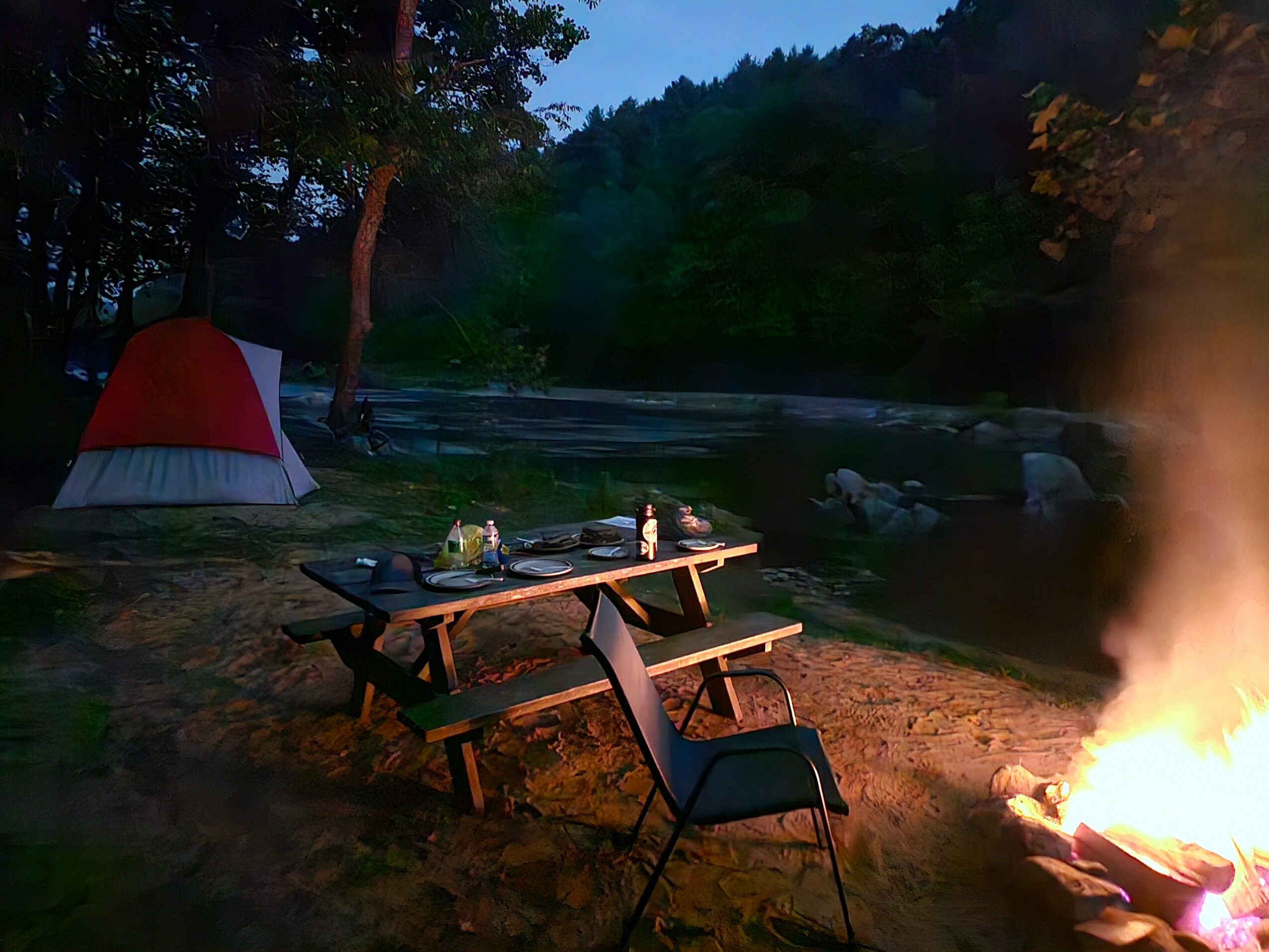 Camping scene with a tent, picnic table, chairs, and a campfire near a river, surrounded by trees at dusk.