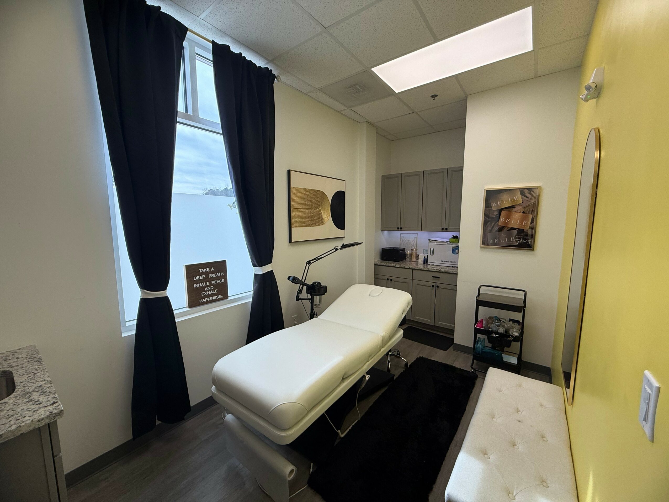 Medical examination room with a bed, window with dark curtains, and artwork on the walls.