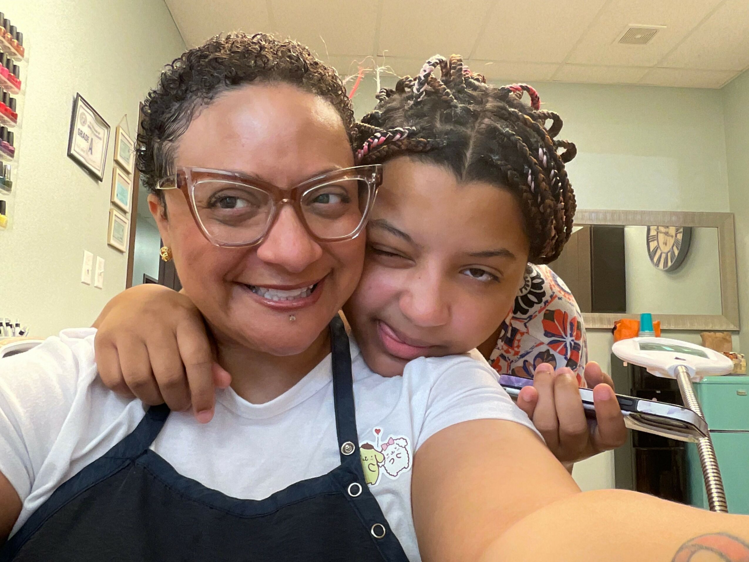 Two people smiling and taking a selfie indoors, one with glasses and curly hair, the other with braided hair, both smiling.