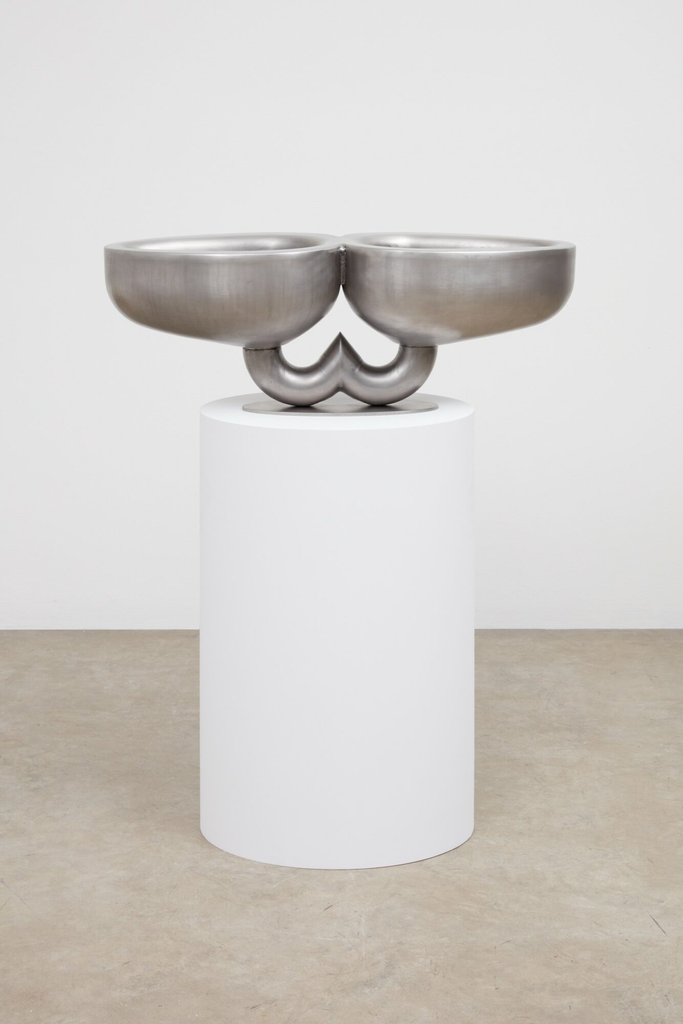 Metal sculpture with two large bowls connected by a heart-shaped element, displayed on a white pedestal against a plain background.