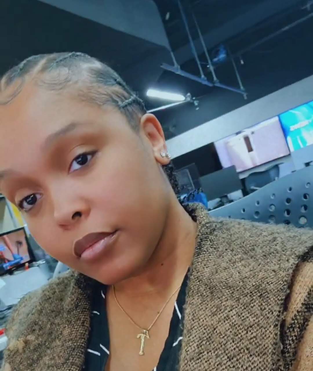 Young woman with braided hair, wearing a gold necklace with a number one pendant, in an indoor setting with screens and equipment.