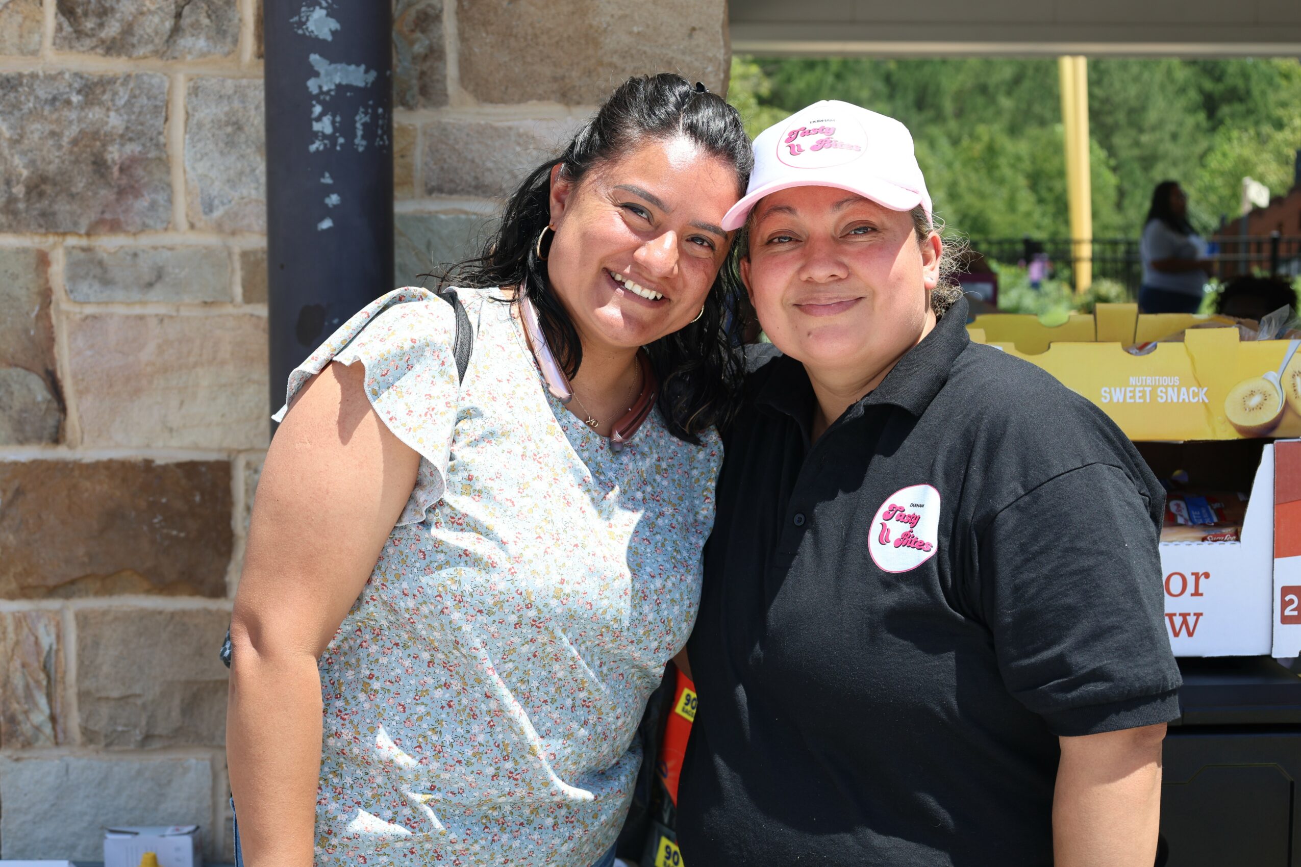 Two women smiling outdoors, one wearing a light-colored top and the other in a black shirt with a white cap, standing close together.