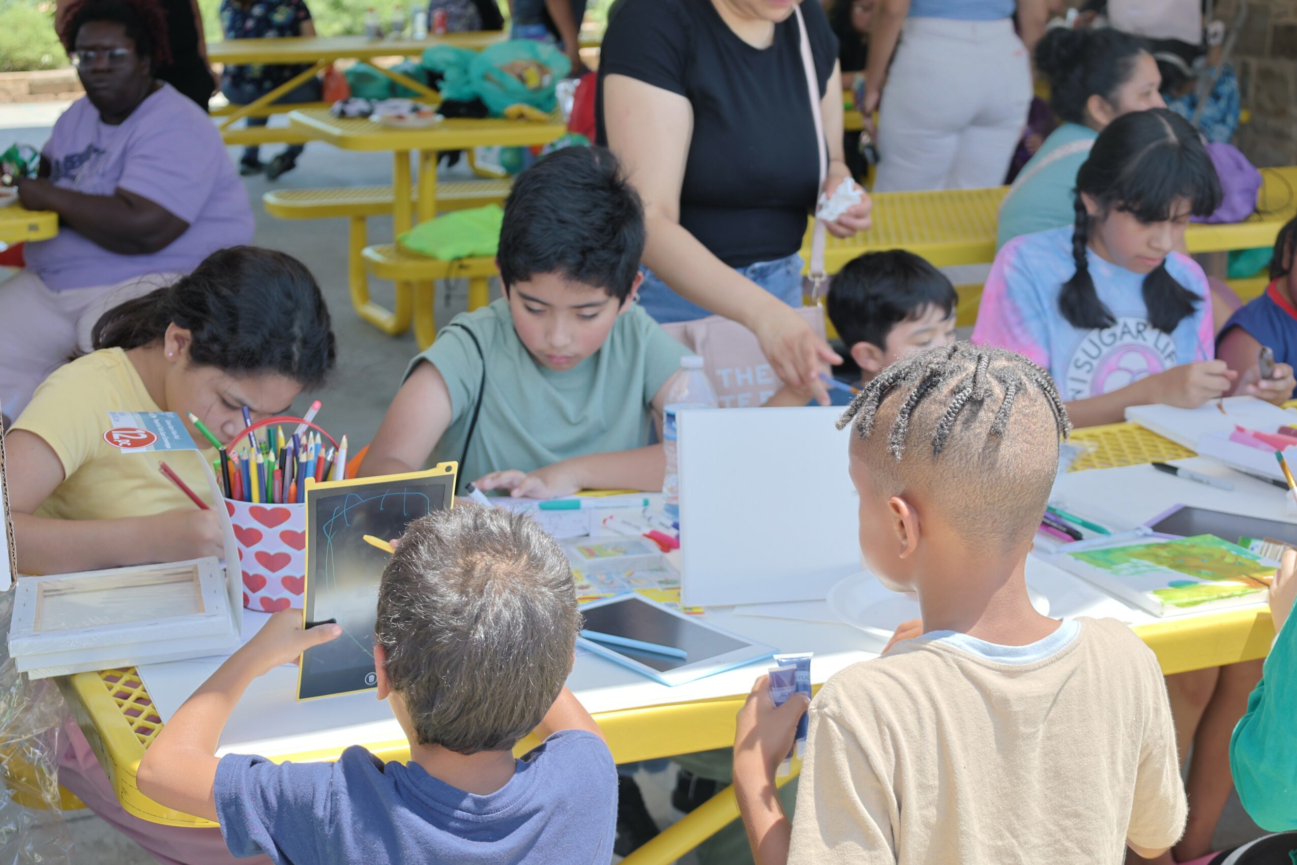 Children sitting at yellow tables drawing and coloring with art supplies, adults supervising, in a bright indoor setting.