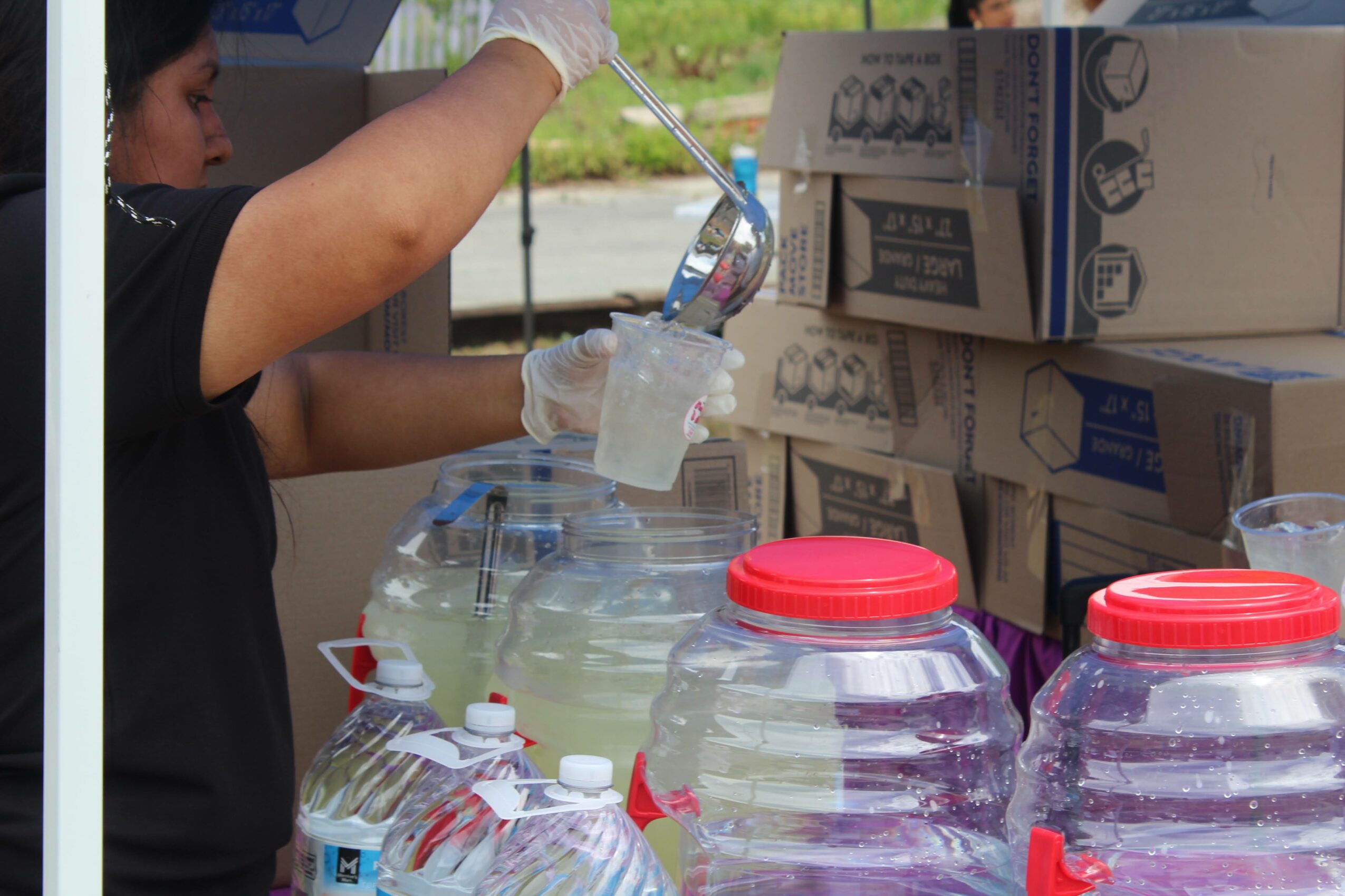 Person scooping liquid from a large container into a cup, with boxes stacked in the background.