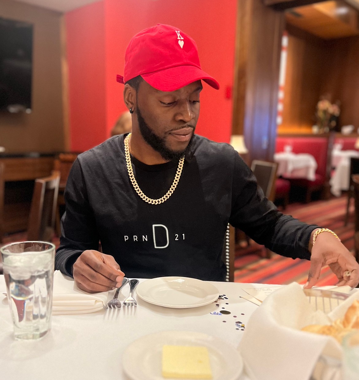 Man wearing a red cap, black shirt, and gold chain sitting at a restaurant table with a glass of water and a plate.