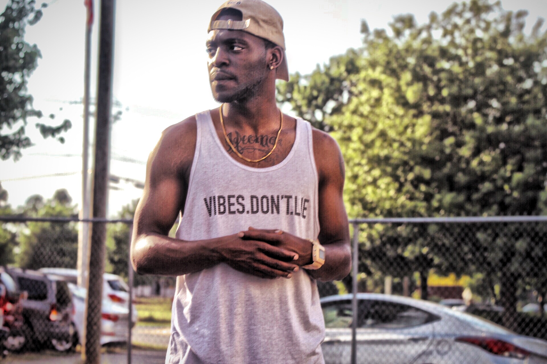 Young man wearing a tank top and cap standing outdoors near a fence with trees and cars in background.