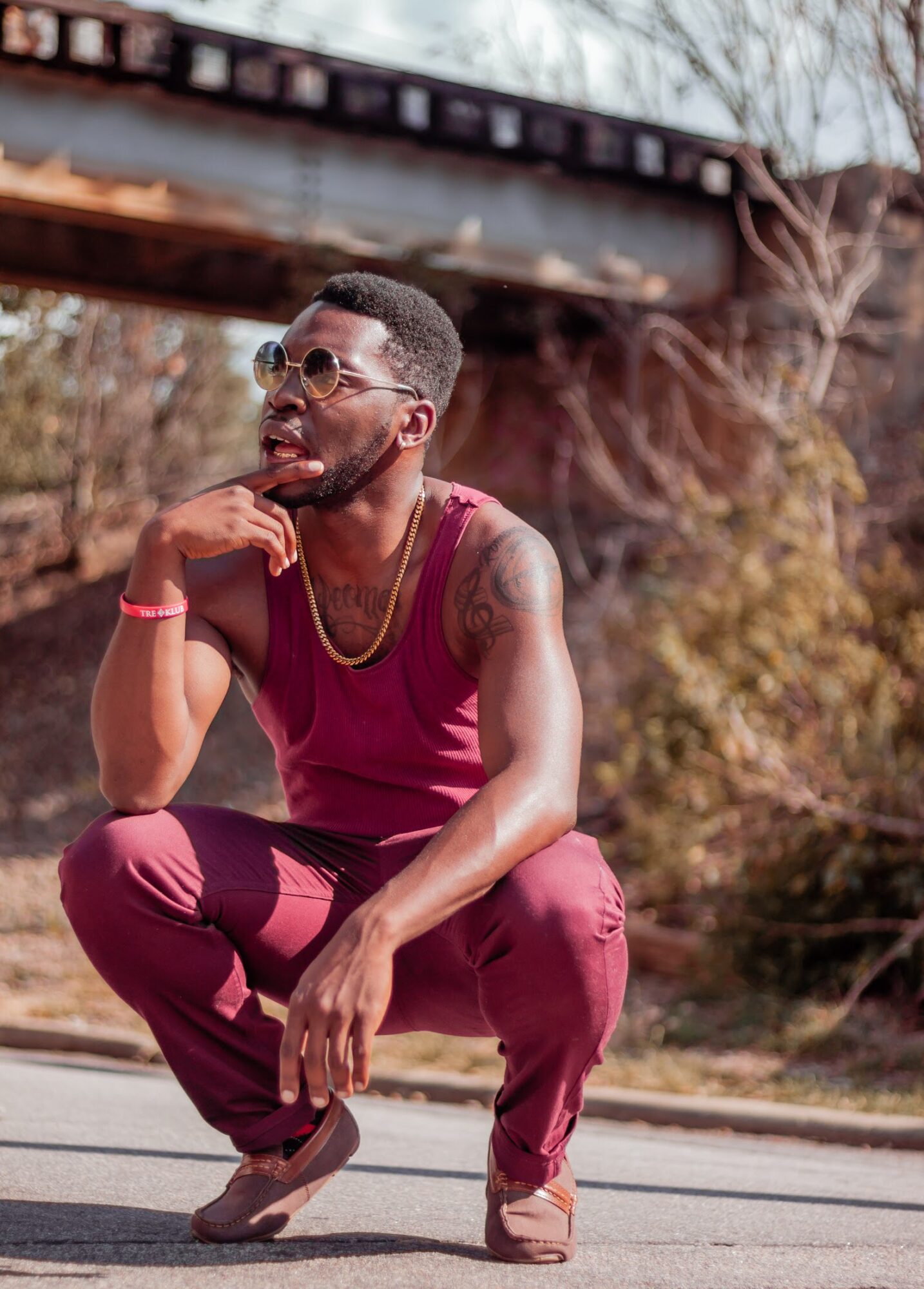 Young man crouching outdoors, wearing sunglasses and a sleeveless maroon outfit, smiling with hand on chin, under a bridge.