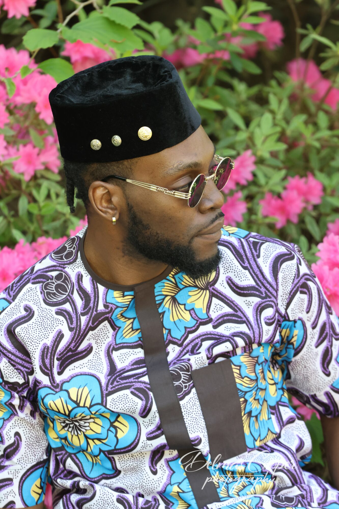 Man wearing traditional attire and sunglasses, standing in front of pink flowers and green foliage.