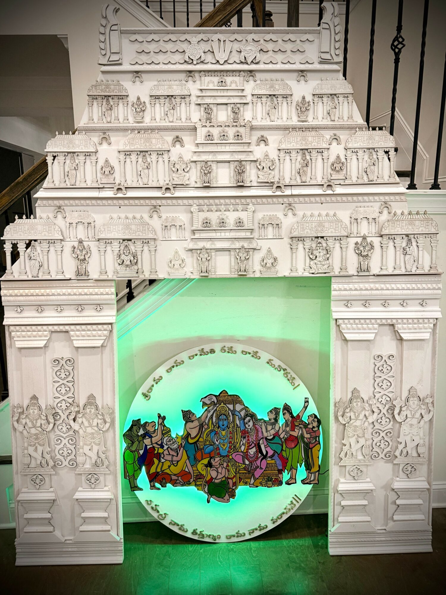 Intricate white carved archway with a circular illuminated scene of colorful deities and figures inside, set against a staircase background.