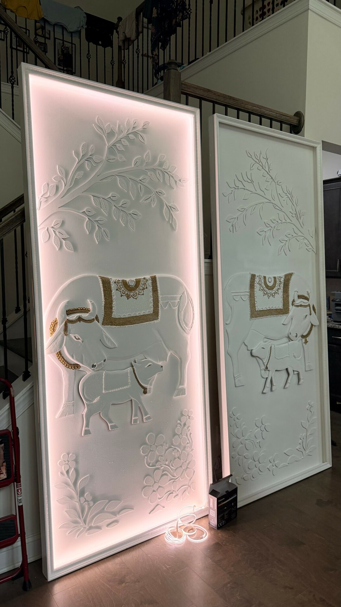 Two decorative panels with elephant and tree designs, one illuminated with pink light, in an indoor setting.