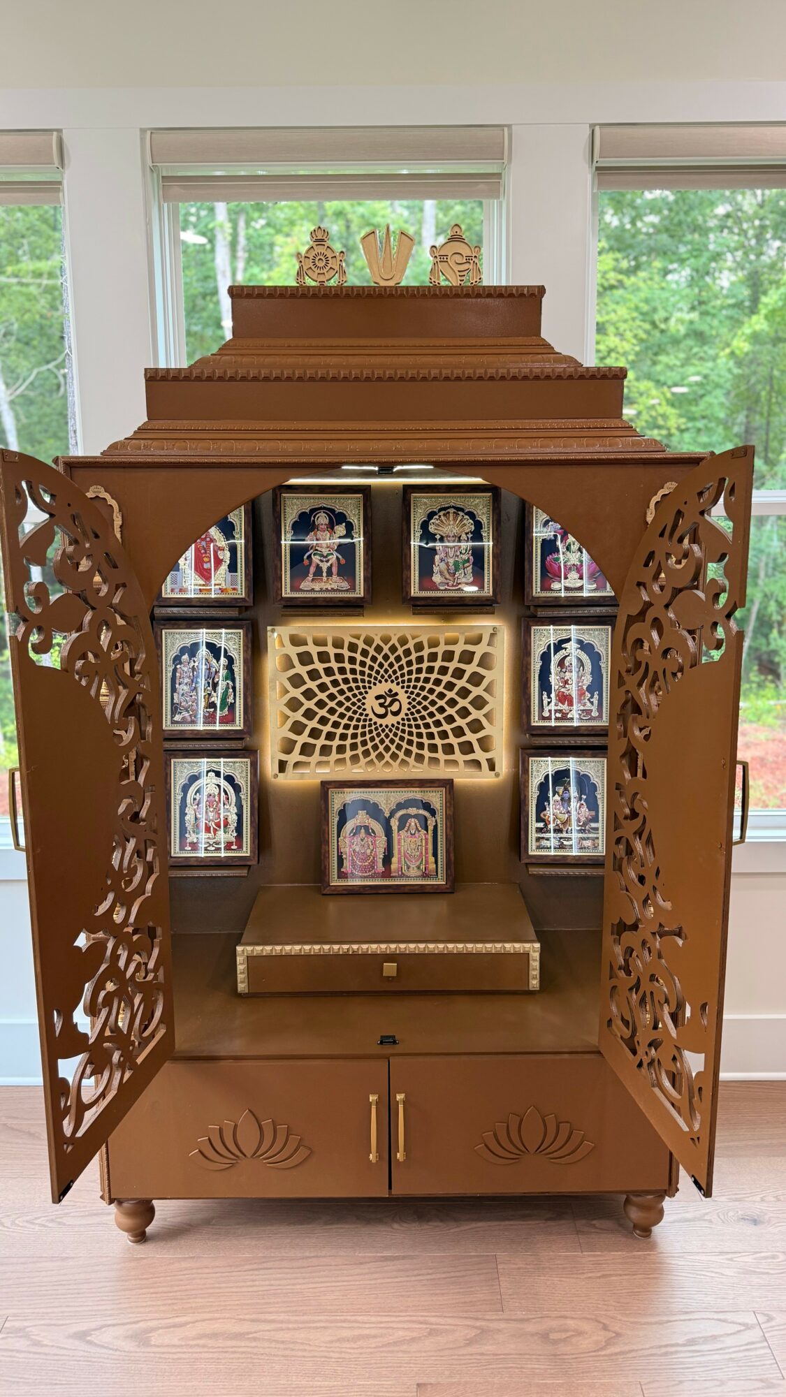 Wooden cabinet with open doors, displaying religious icons and decorative elements, set in a room with large windows and greenery outside.