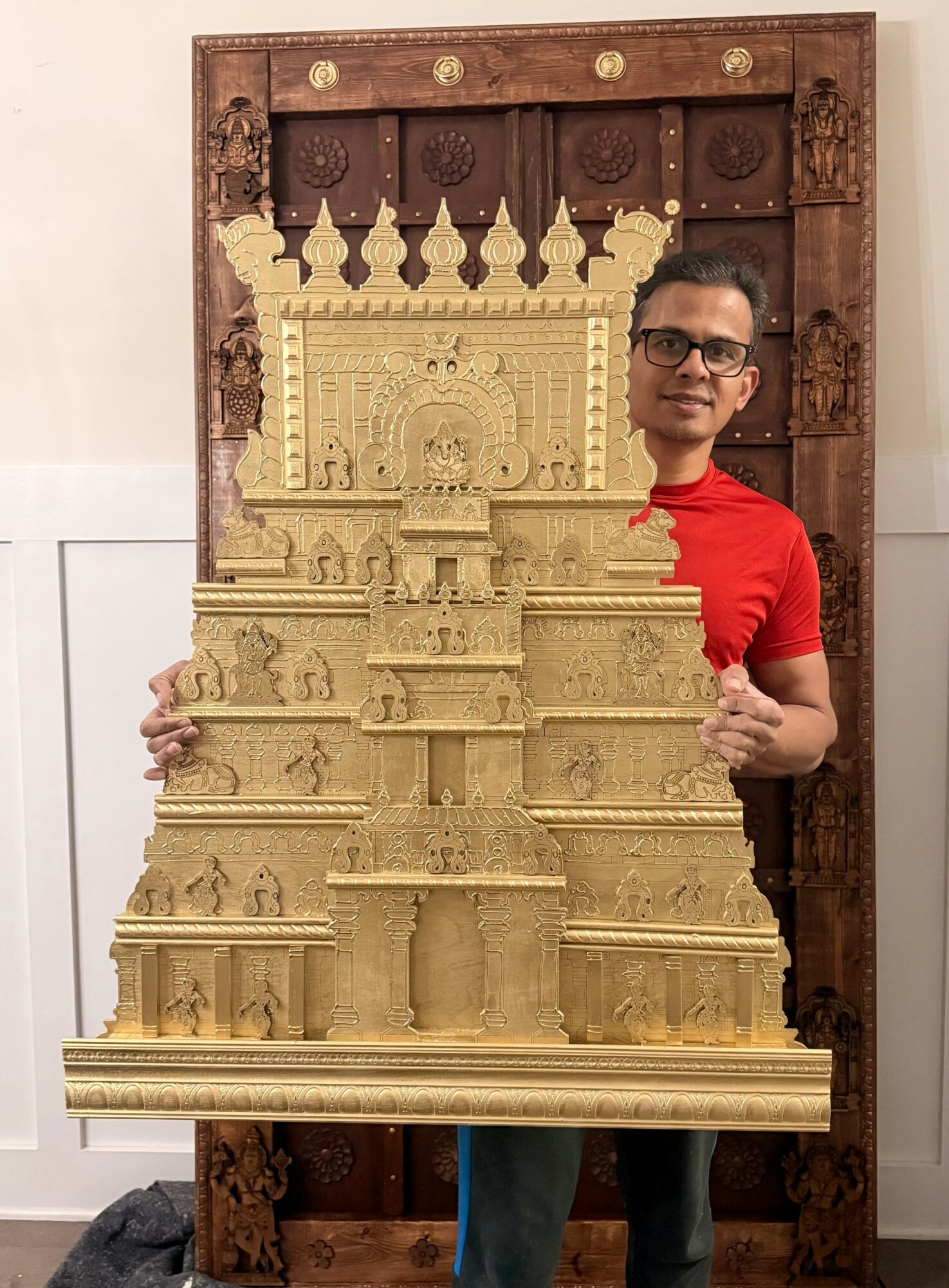 Person holding a large, detailed carved wooden structure, standing in front of a wooden frame, wearing glasses and a red shirt.
