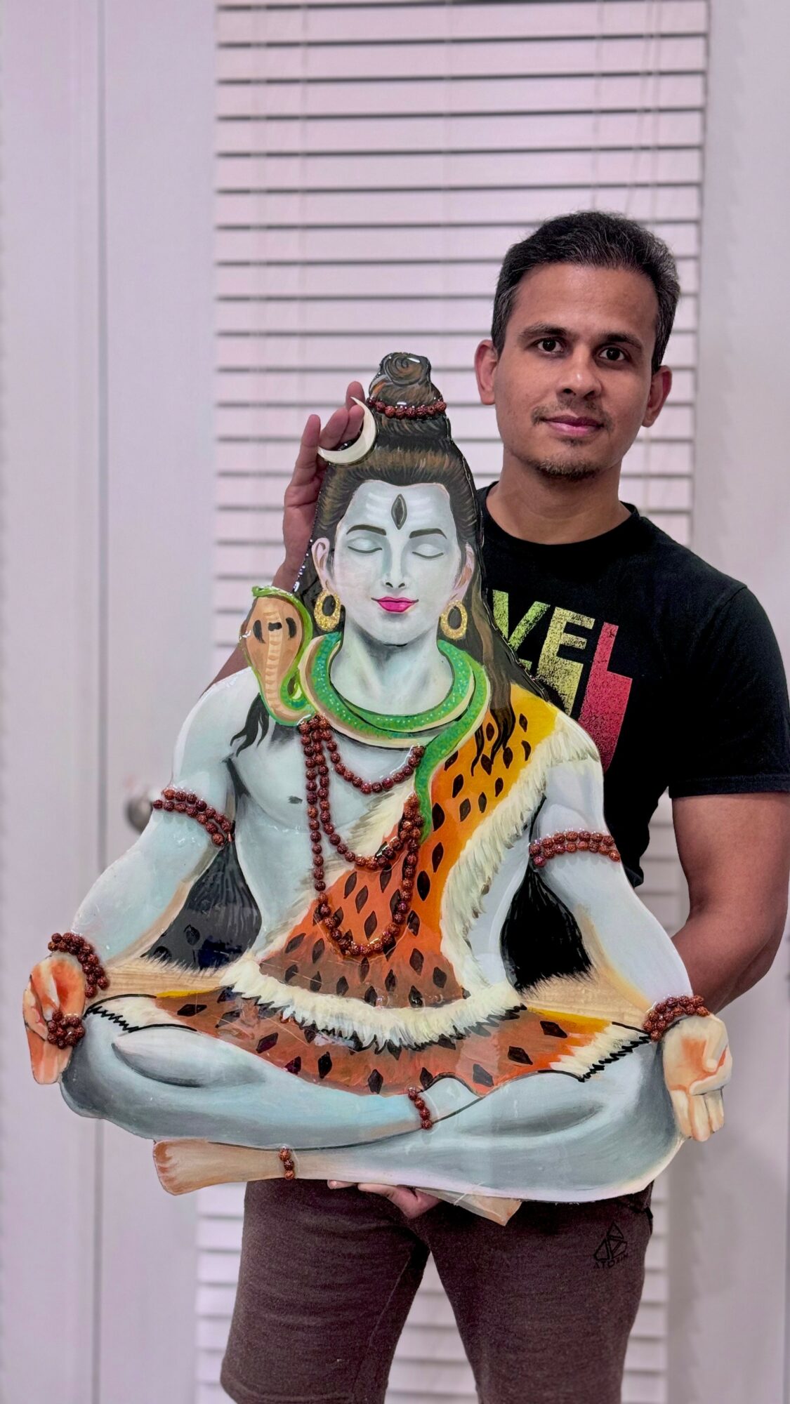Man standing behind a statue of a Hindu deity, with a white background and closed blinds.