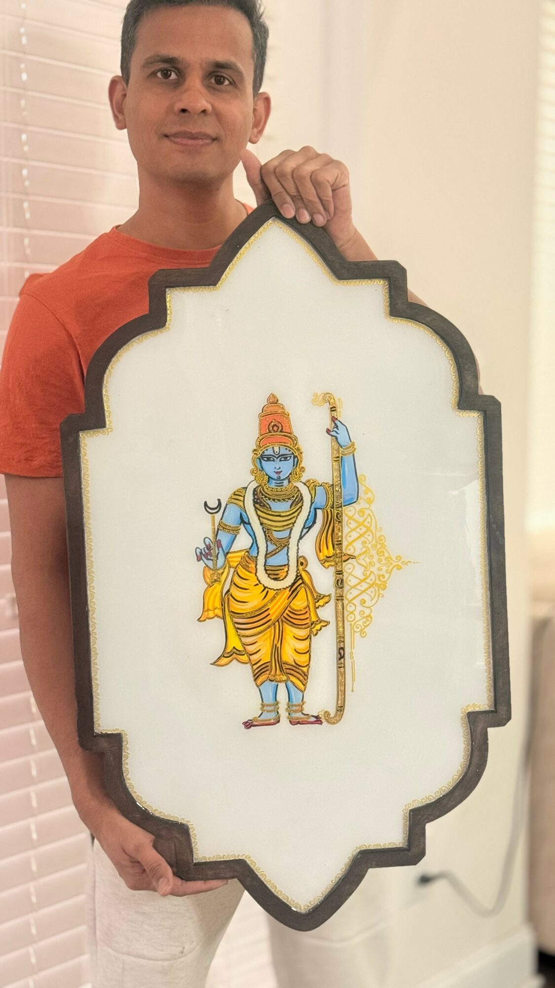 Person holding a framed picture of a Hindu deity, standing indoors with blinds in the background.