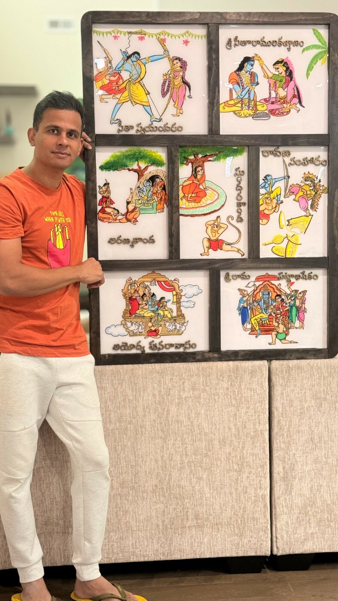 Man standing next to a display of eight colorful traditional paintings on a black board, with a beige background.