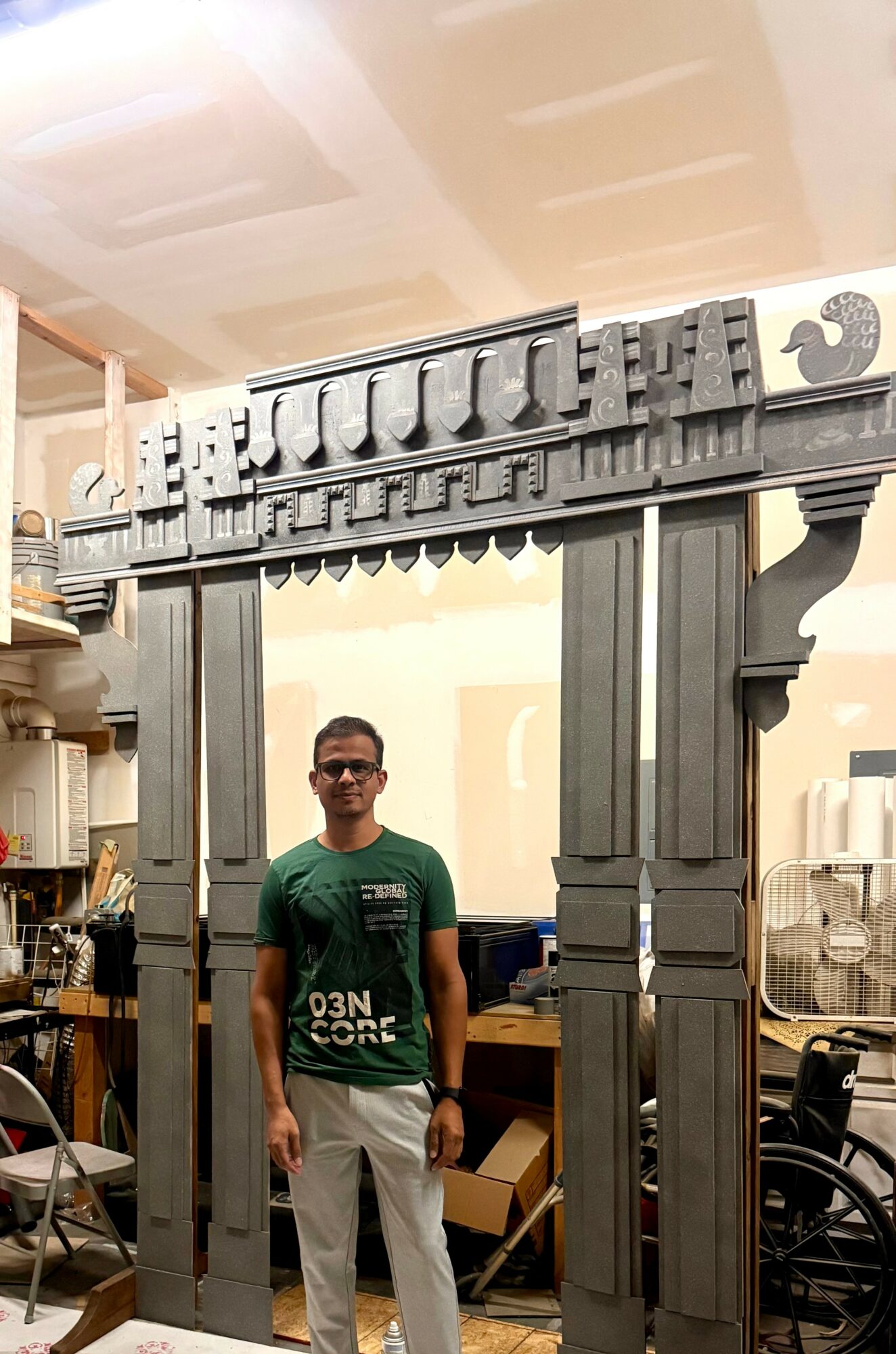 Man wearing sunglasses and green shirt standing in front of a large decorative structure, indoors.