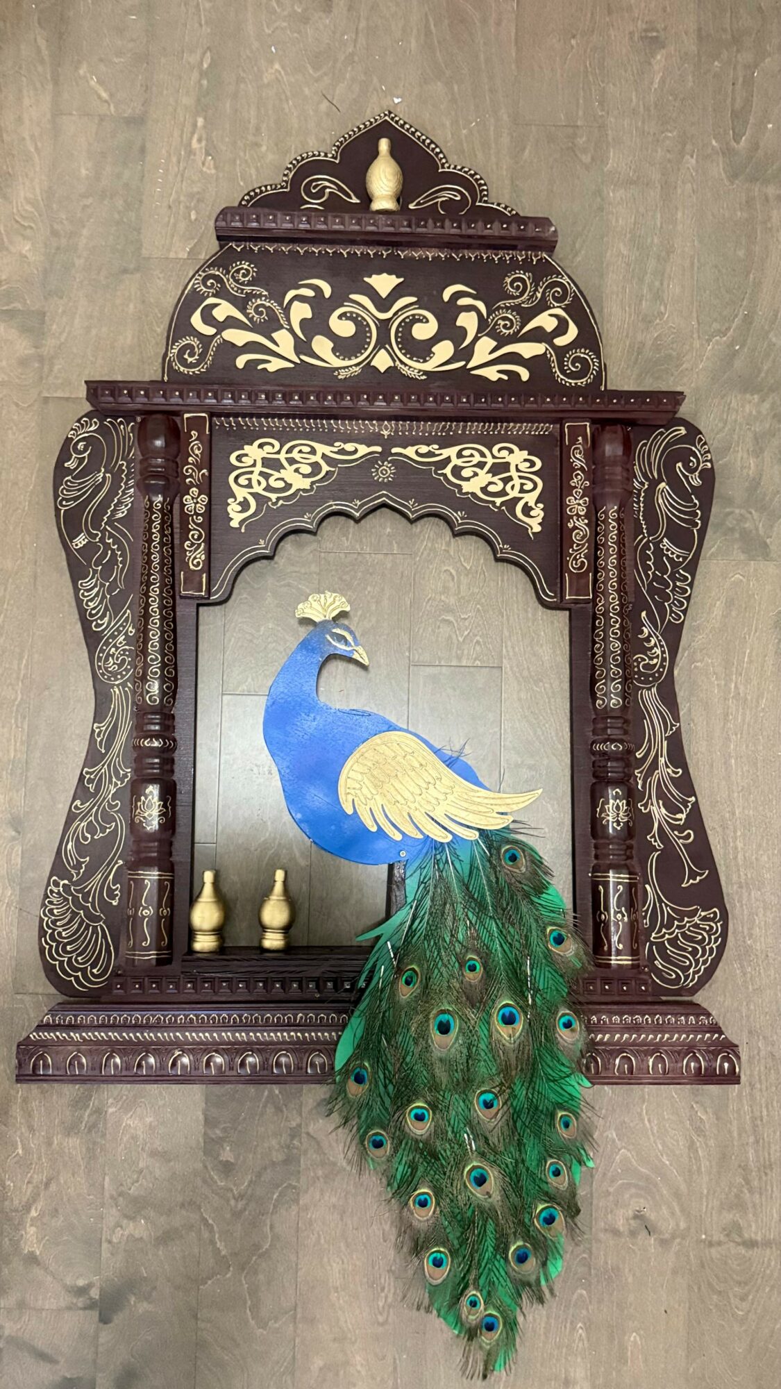 Decorative wooden frame with a peacock illustration and real peacock feathers hanging below, set against a gray background.