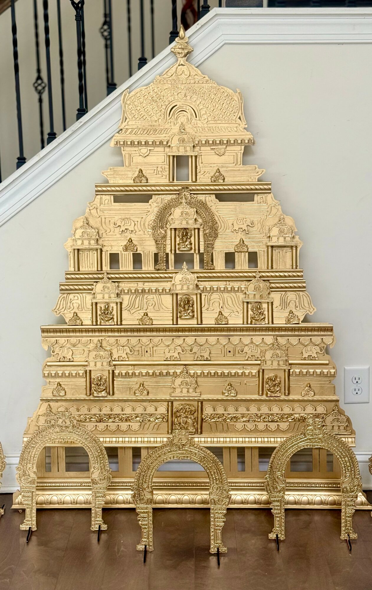 Golden temple structure with multiple tiers, arches, and intricate carvings, placed against a white wall and staircase, on a wooden floor.