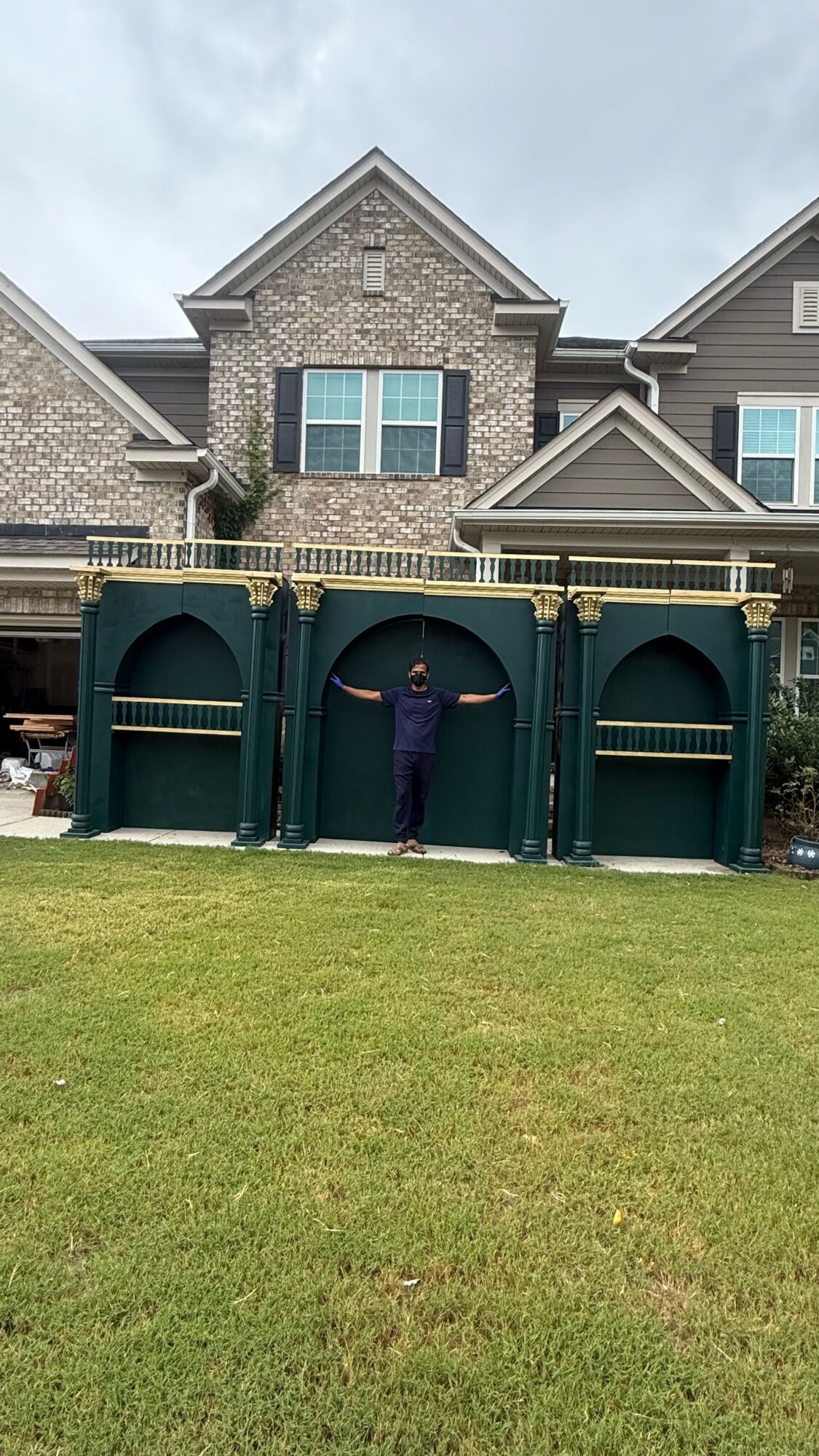 Person standing in front of a green decorative fence with arches, in front of a brick house with multiple windows and a gabled roof.