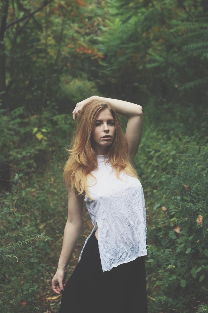 Young woman with long hair standing outdoors in a green forest, wearing a white sleeveless top and black skirt, looking at the camera.