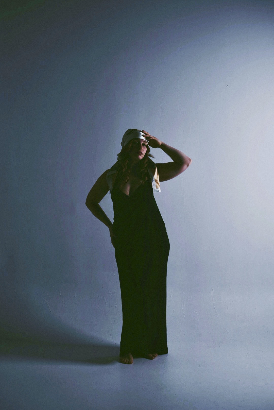 Person standing in a dark outfit, holding sunglasses, with a gradient background from dark to light.
