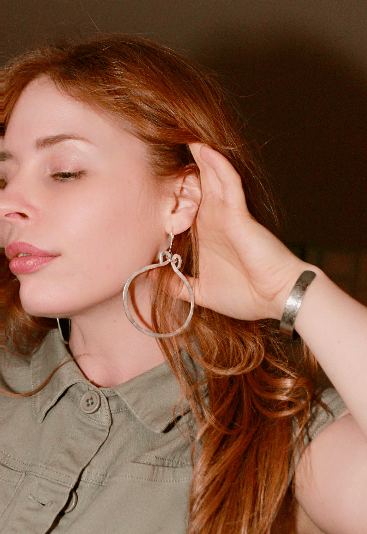 Woman with red hair and hoop earrings, eyes closed, touching her hair, wearing a gray shirt, indoors.