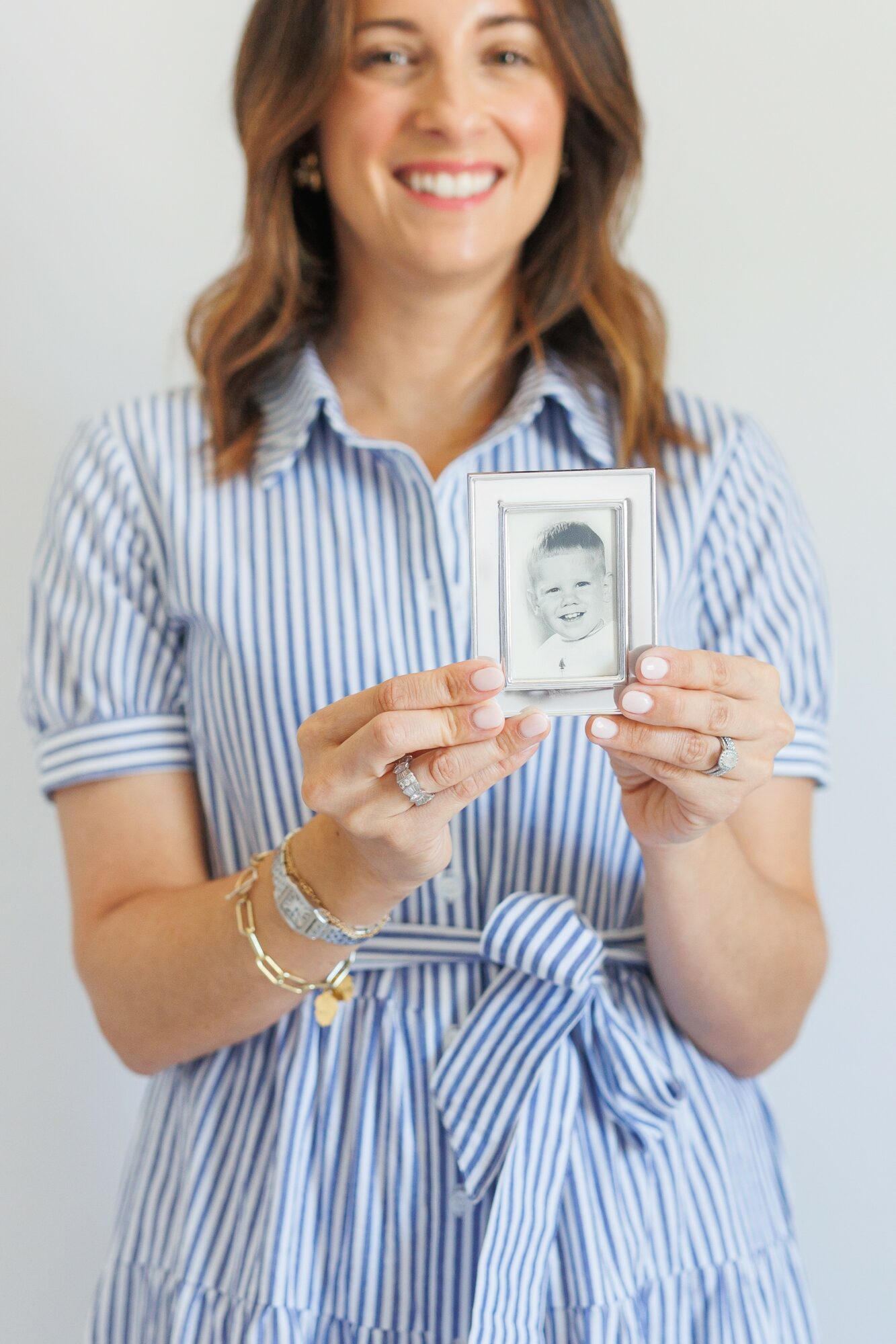 Woman smiling holding a black-and-white photo of a person, wearing a striped dress, against a plain background.