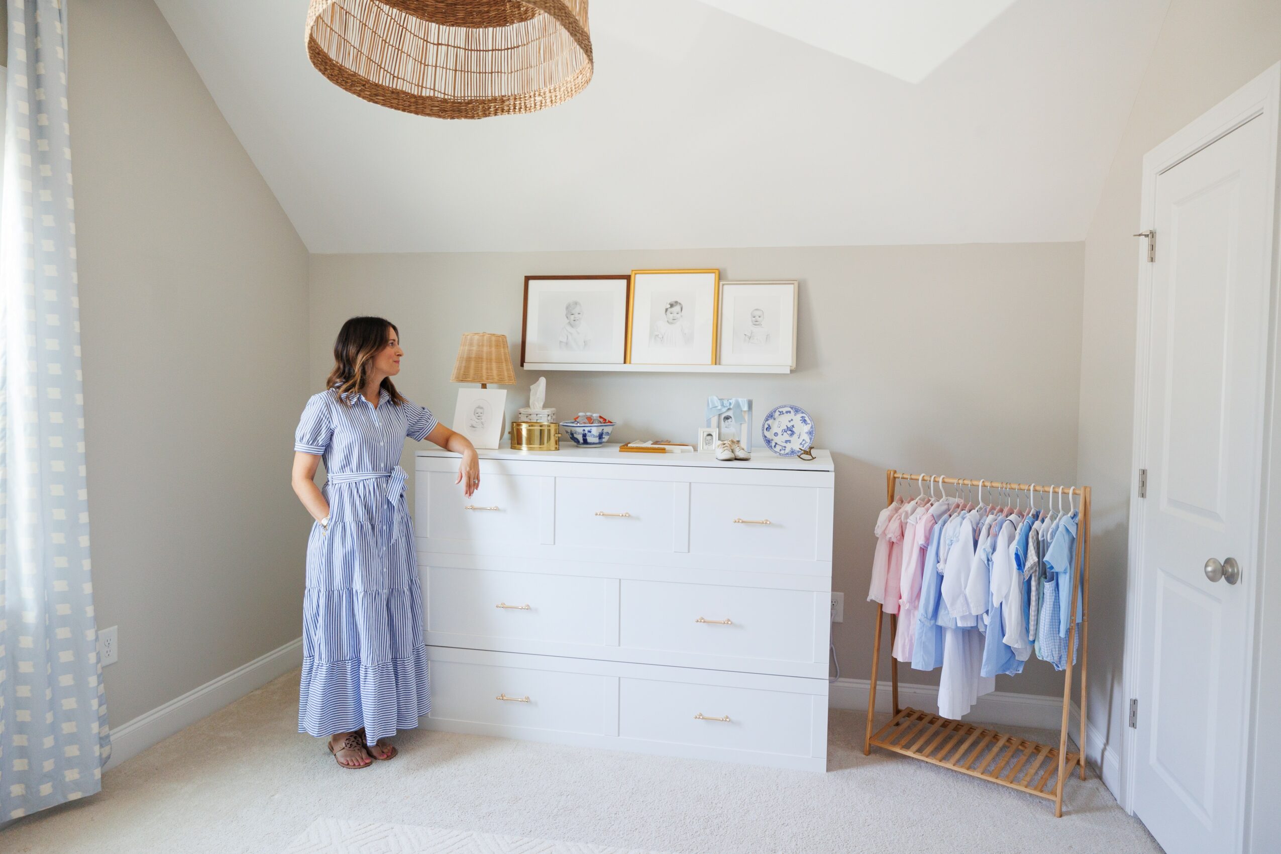 Woman in a blue and white striped dress standing in a light-colored bedroom with a white dresser, framed artwork, and a clothing rack.