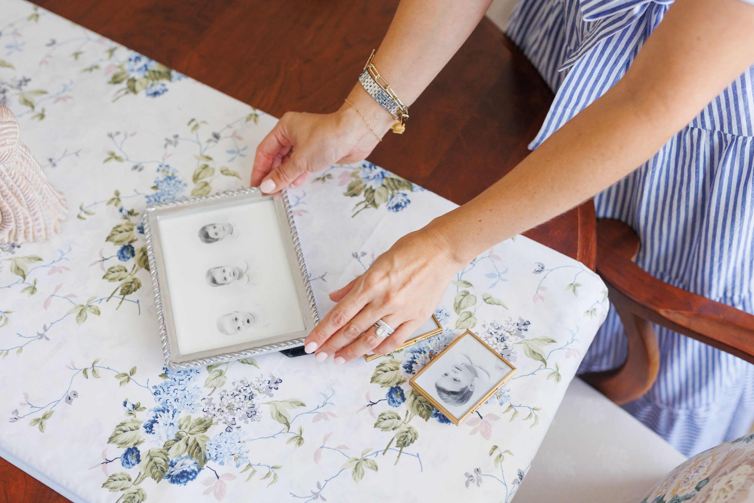 Person's hands arranging jewelry on a floral tablecloth with a jewelry box and a small framed picture nearby.