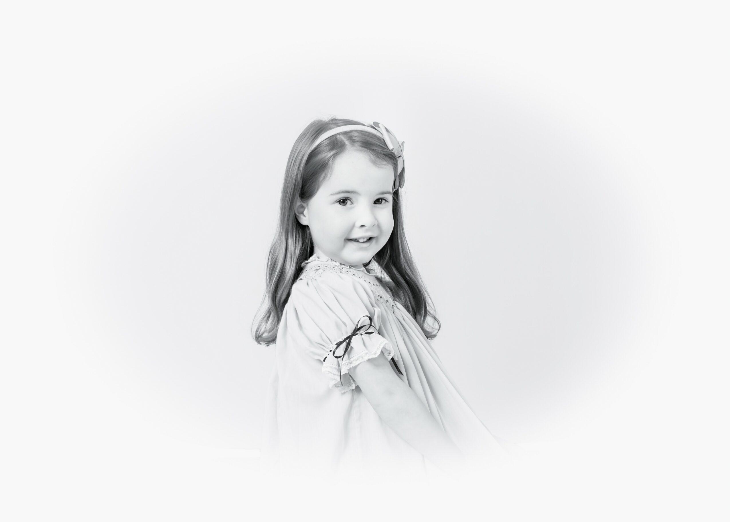Young girl with long hair and a bow, smiling, wearing a dress, facing slightly to the right, in black and white.