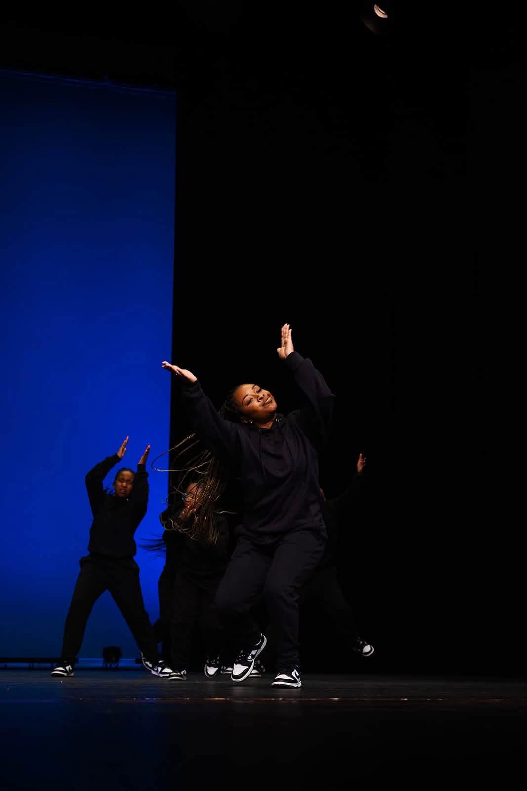Two dancers perform on stage, one in front with arms raised, the other behind with one arm up, against a blue background.