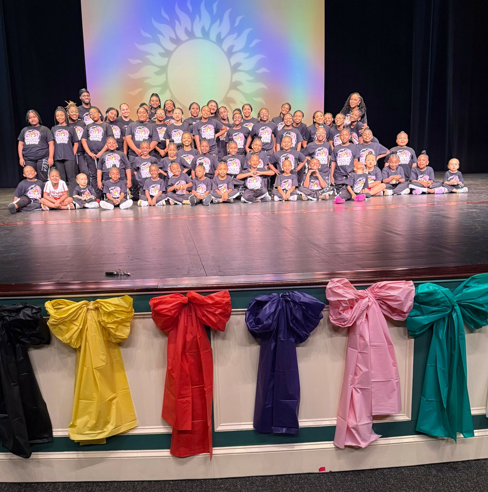 Group of children and adults on stage, with colorful decorations and a projected sun design in the background.