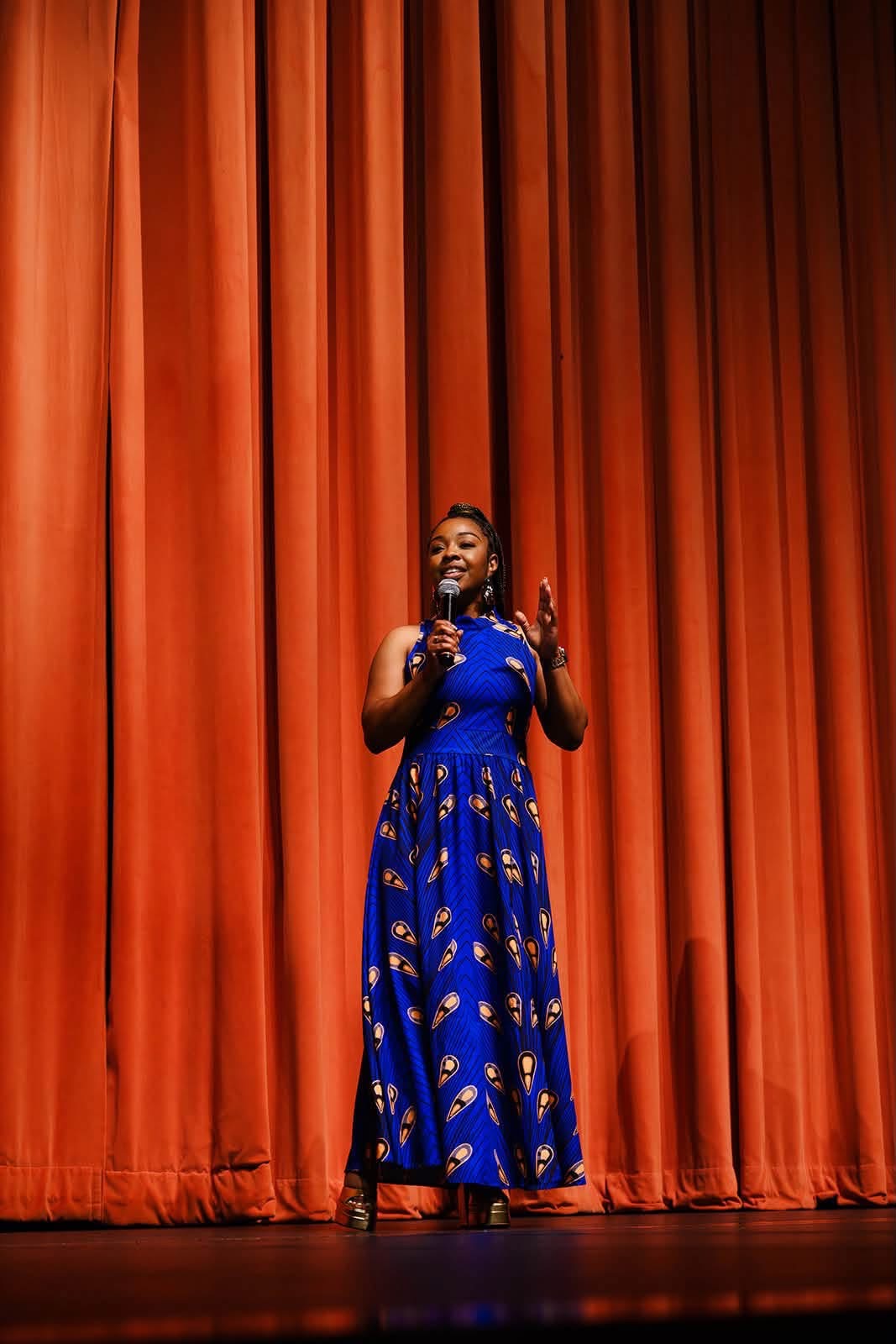 Woman in blue dress standing on stage with orange curtains behind her, speaking or singing, holding a microphone.