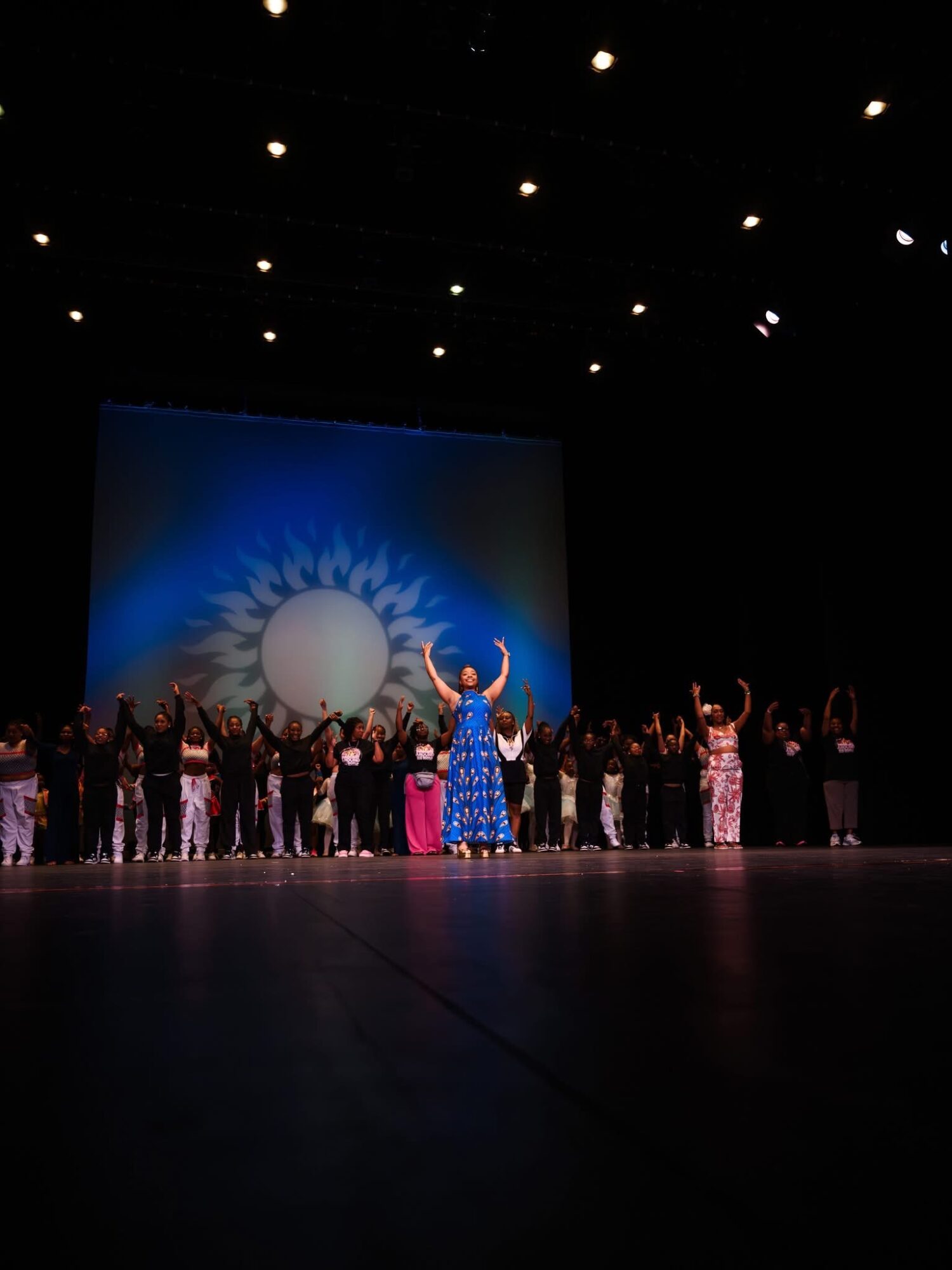 Stage performance with a group of people, a woman in a blue dress in front, and a backdrop with a sun and shadow pattern.
