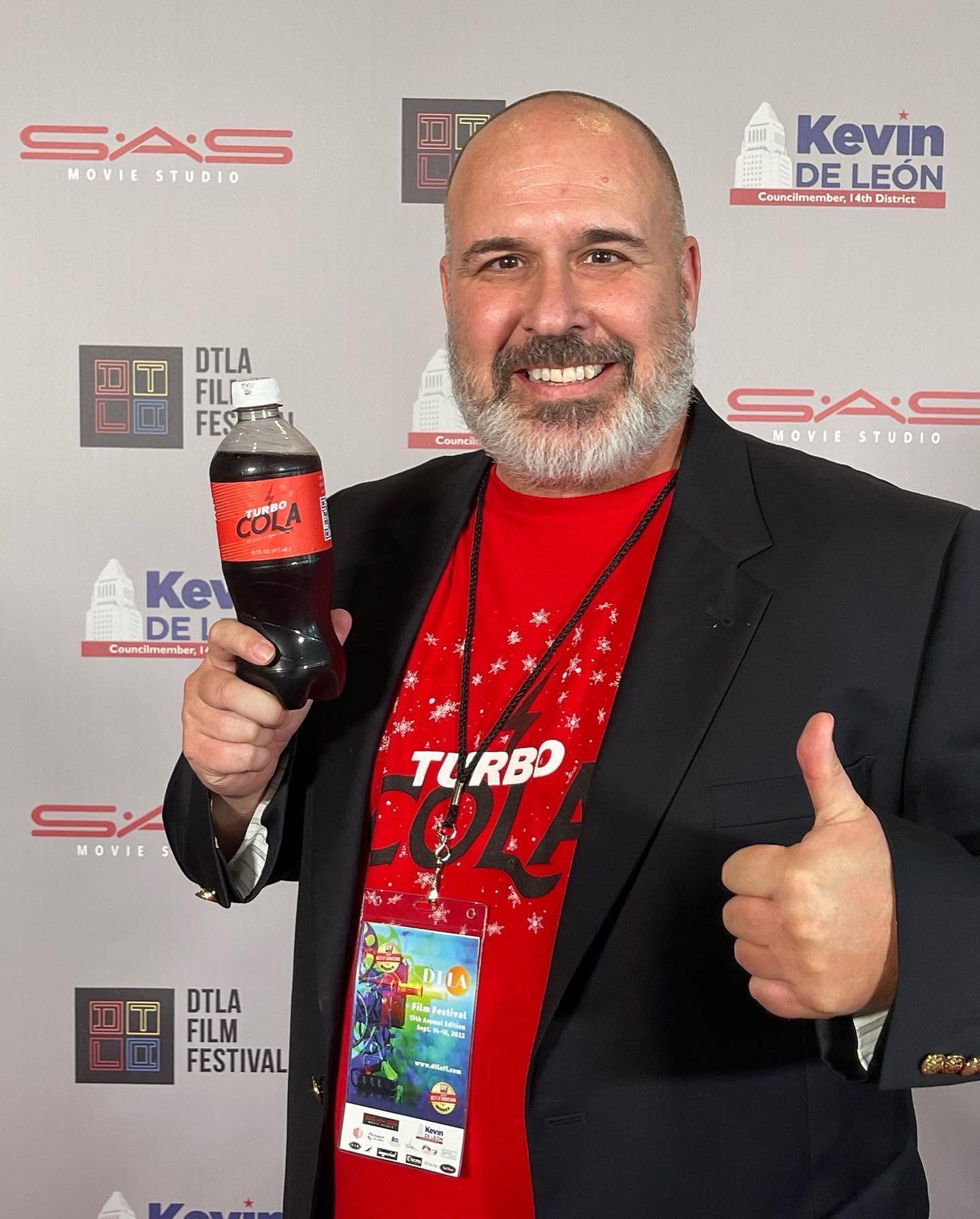 Man with a beard in a suit holding a soda bottle and giving a thumbs-up, standing in front of a festival backdrop.