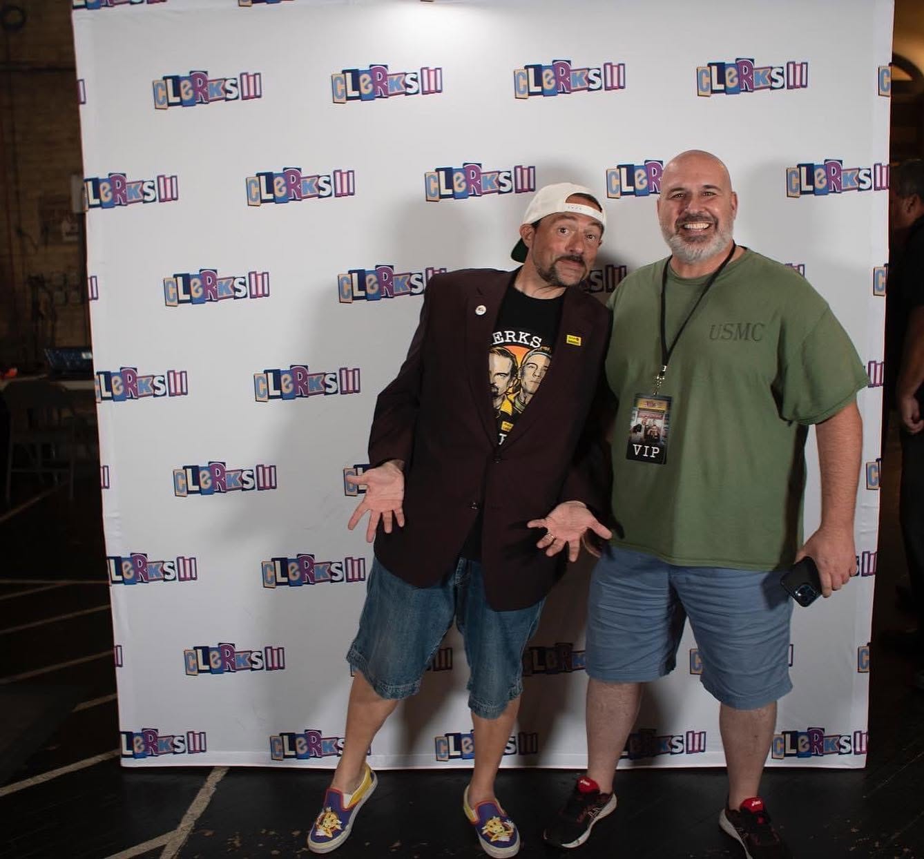 Two men stand in front of a backdrop with a logo, smiling and posing for a photo, one wearing a hat and shorts.